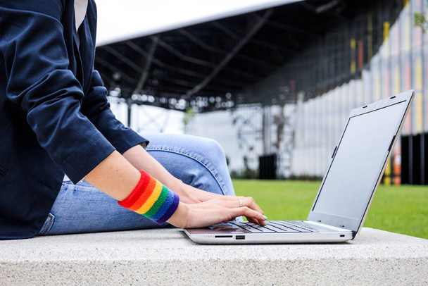 Női kezek közelsége laptoppal és LMBT karkötővel a modern parkban. LMBTQIA kultúra. - Fotó, kép