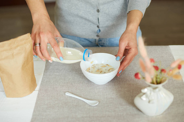 Frau bereitet gesundes Frühstück zu Hause zu, weibliche Hand hält Flasche, die Milch in Müsli-Flocken-Schüssel mit Nüssen Rosinen, Müsli-Essen Hafermehl, Lifestyle-Konzept - Foto, Bild