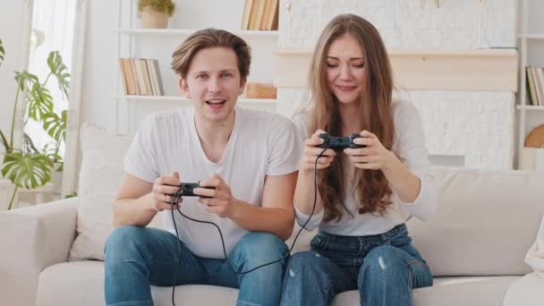Beyaz tenli evli çift Millennial erkek ve erkek arkadaş, kanepede oturmuş video oyunu oynuyorlar. Konsol kontrolörleri ile yarışan kız zafer jesti yapıyor. - Video, Çekim