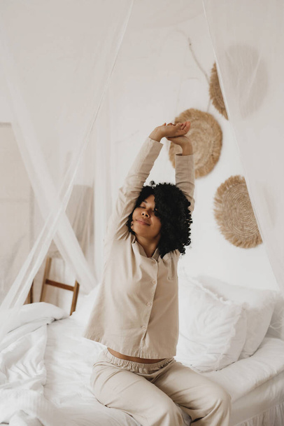 African American woman in sleepwear stretching and smiling with closed eyes sitting on bed at home. - Foto, afbeelding