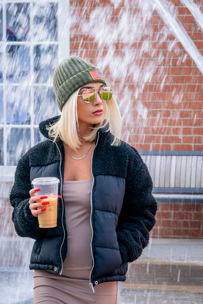 Ein hinreißendes junges blondes Model posiert im Freien, während es einen Wintertag in einer städtischen Umgebung genießt und ihren Kaffee trinkt - Foto, Bild