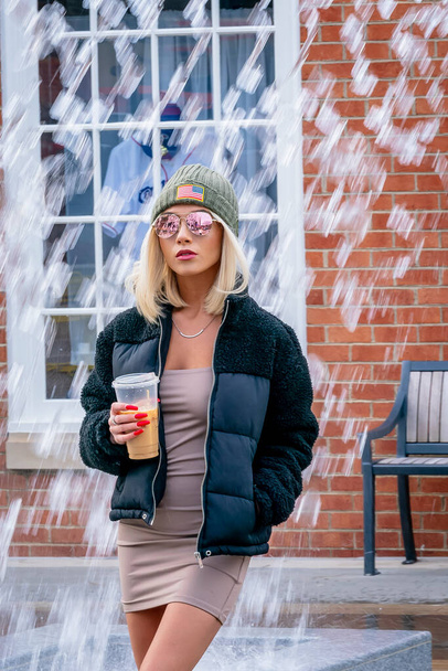 A gorgeous young blonde model poses outdoors while enjoying a winters day in a city environment drinking her coffee - Photo, image