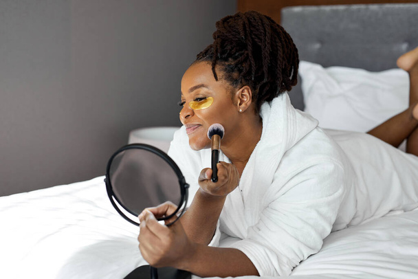 φροντίδα ομορφιάς στο σπίτι. Ευτυχισμένη Αφρικανή γυναίκα με μπουρνούζι μετά το ντους που εφαρμόζει αντικόπωση κάτω από τη μάσκα ματιών και μακιγιάρεται - Φωτογραφία, εικόνα