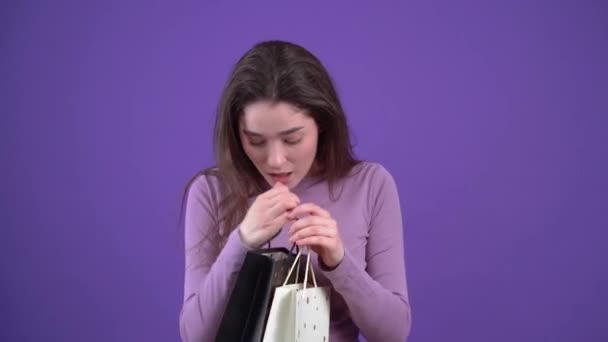 The curious girl looks at the package and is pleasantly surprised by the gift she received. Brunette isolated on a purple background, dressed in a purple sweater. Lifestyle concept - Video, Çekim