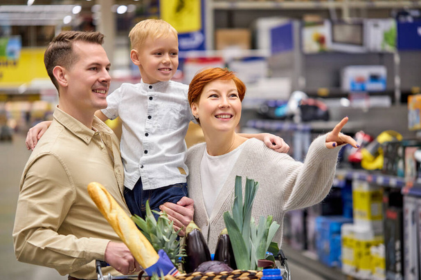 Χαμογελαστή οικογένεια αγκαλιάζει, χαμογελά, ερωτεύεται ενώ ψωνίζει σε σούπερ μάρκετ στέκεται σε εσωτερικούς διαδρόμους καταστημάτων - Φωτογραφία, εικόνα
