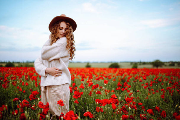 Beautiful girl posing in a poppy field. Young woman stands in a spring blooming garden. A curly-haired girl stands in blooming field in summer. Sunny day, summer landscape. Joy and relaxation concept - Foto, Imagem