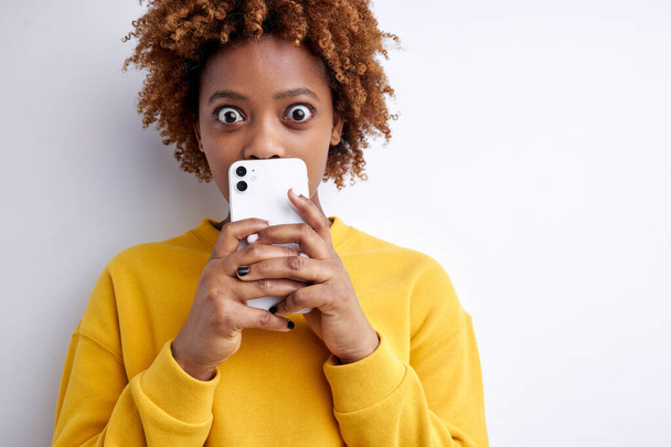 afro american female is very surprised and shocked by what she sees on mobile phone, look camera with wide opened eyes - 写真・画像