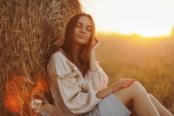 fiatal nő a gyönyörű fényében a nyári naplemente egy mezőn ül a szalma bálák. gyönyörű romantikus lány hosszú haj szabadban mező. - Fotó, kép