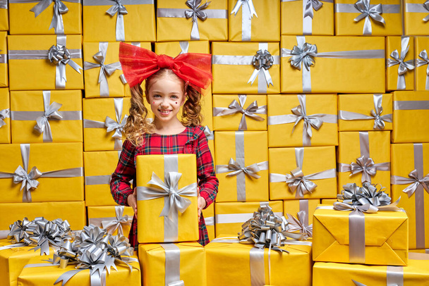 Excited caucasian kid girl in checkered red dress among many gifts. International Womens Day birthday, holiday concept - 写真・画像