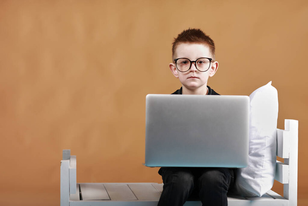 Egy szemüveges iskolás srác laptoppal ül az ágyon. Gyermek csinál iskolai házi feladatot, és komolyan és fáradtan néz kamera.Online tanulás, távoktatás, oktatás online oktató otthon elszigeteltség. - Fotó, kép