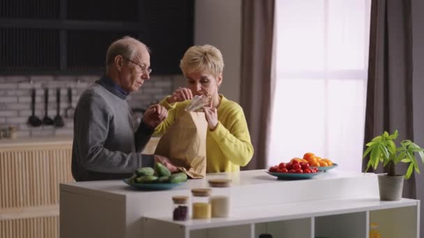 pareja de ancianos casados está desempacando bolsa con comida en la cocina casera, par de jubilados está poniendo comida en la mesa, retrato medio en el apartamento - Metraje, vídeo
