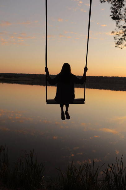 silhouet van een romantische jonge vrouw op een schommel boven meer bij zonsondergang. jong meisje reiziger zitten op de schommel in prachtige natuur, uitzicht op het meer. - Foto, afbeelding