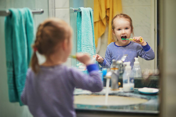 Happy toddler girl in pyjamas brushing her teeth in bathroom in the morning or before going to sleep. Dental hygiene for small kids - Foto, Imagen