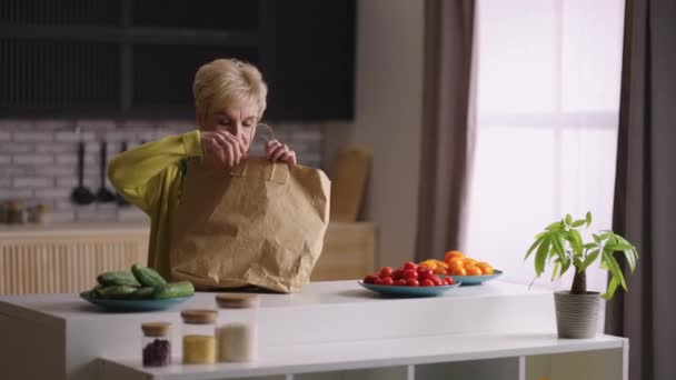 Yaşlı kadın süpermarketten dönüyor ve yiyecek dolu çantalarını açıyor, mutfak masasına eşya koyuyor. - Video, Çekim