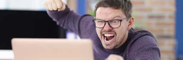 Wütender Mann mit Brille fuchtelt mit der Faust auf Laptop-Monitor - Foto, Bild