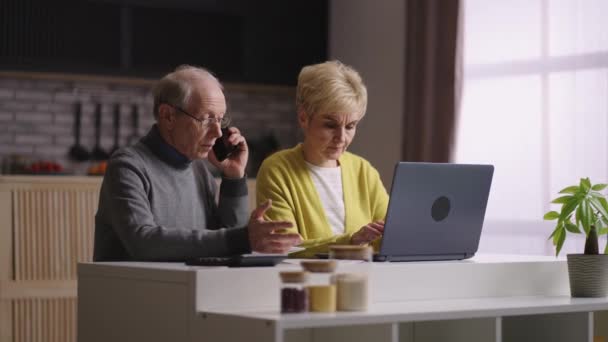 starý muž a žena platí účty za služby on-line, pobyt doma, manžel volá po telefonu a žena používá notebook s internetem - Záběry, video