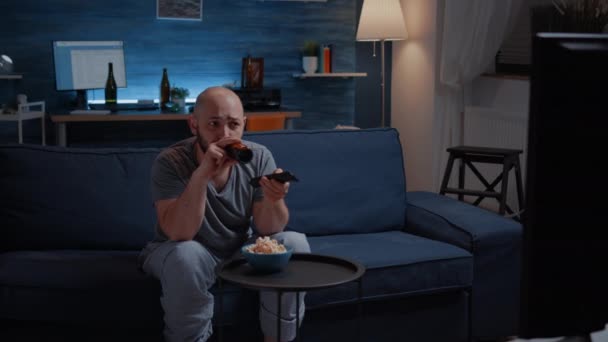 Gelangweilter Mann, der Fernsehsender wechselt, sitzt auf gemütlicher Couch und trinkt Bier - Filmmaterial, Video