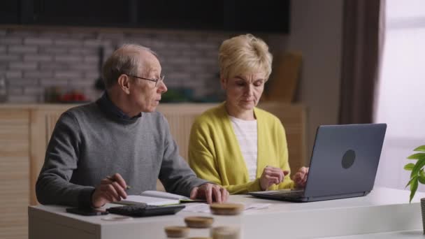 důchodci muž a žena jsou výpočet výdajů sedí společně v domácí kuchyni, starý muž je počítání podle kalkulačky a stará dáma používá notebook - Záběry, video