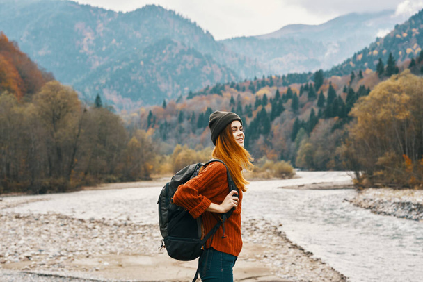 радостная женщина туристка с рюкзаком смотрит на горы у реки на берегу - Фото, изображение