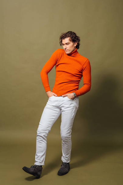 Ganzkörperporträt eines großen, gut aussehenden Mannes mit orangefarbenem Rollkragen und weißen Jeans, der auf grünem Hintergrund posiert - Foto, Bild