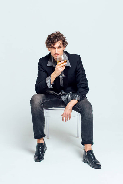 Tall handsome confident young man in a jacket sitting on the chair and holding glass of wine over white background - Fotografie, Obrázek
