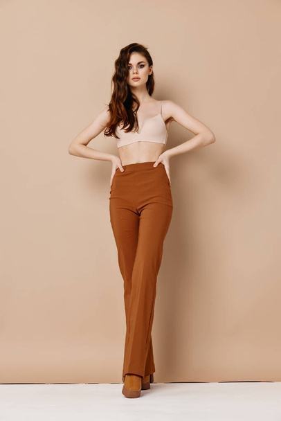 an extravagant woman in trousers and a bra on a beige background holds her hands on her belt - Photo, Image