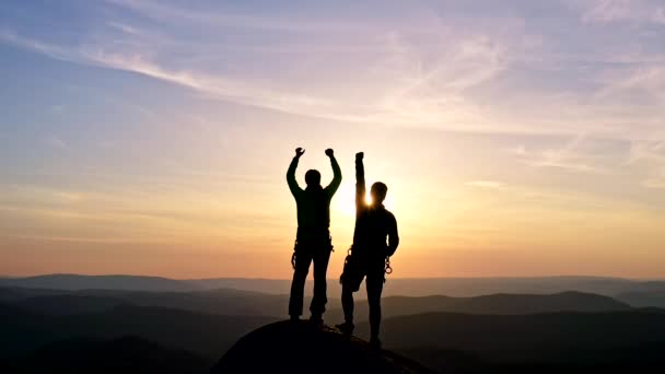 Egy boldog ifjú pár sziluettjei egy hegy tetején, akik diadalmasan emelik fel a kezüket a levegőben. - Felvétel, videó