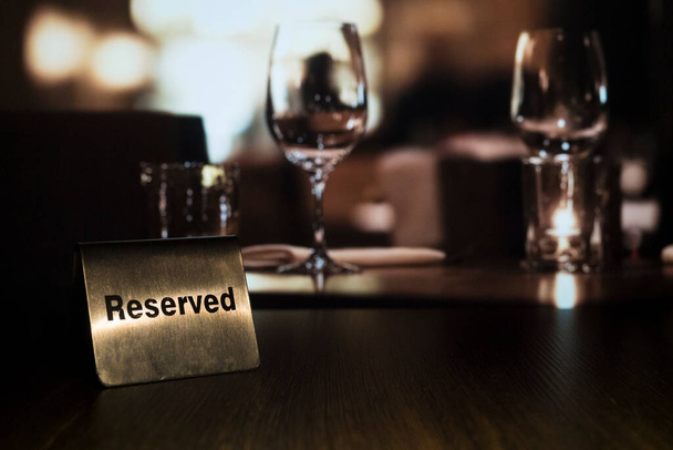 Egy fém emléktábla a "Reserved" felirattal baloldalt áll egy fa asztalon egy étteremben, gyertyával és szemüveggel a háttérben.. - Fotó, kép
