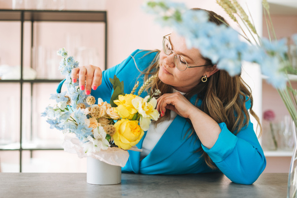 Νεαρή γυναίκα ανθοπώλης σε φωτεινό μπλε κοστούμι και γυαλιά με μπουκέτο λουλούδια σε κουτί σε ανθοπωλείο, μικρό τοπικό ιδιοκτήτη της επιχείρησης. Νεαρή κομψή επιτυχία χιλιετή γυναίκα στη δημιουργική εργασία - Φωτογραφία, εικόνα