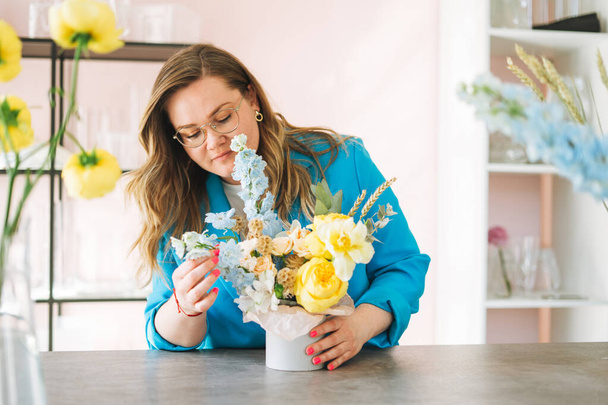 Młoda kobieta kwiaciarnia w jasnoniebieskim garniturze i okularach oko z bukietem kwiatów w pudełku w kwiaciarni, mały lokalny właściciel firmy. Młoda stylowa kobieta sukcesu tysiąclecia na twórczej pracy - Zdjęcie, obraz