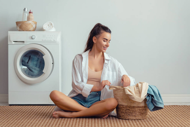 Beautiful young woman is smiling while doing laundry at home. - Foto, imagen