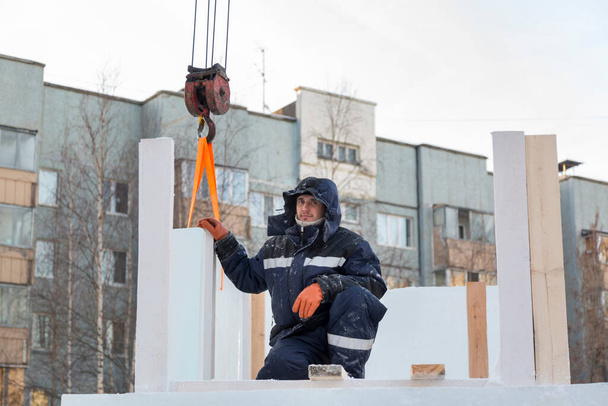 Портрет рабочего в синей куртке с капюшоном на крючке грузового крана на строительстве ледового городка - Фото, изображение