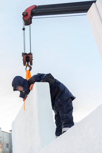 Retrato de un trabajador con una chaqueta azul con una capucha en el gancho de una grúa de camión en la construcción de una ciudad de hielo - Foto, Imagen