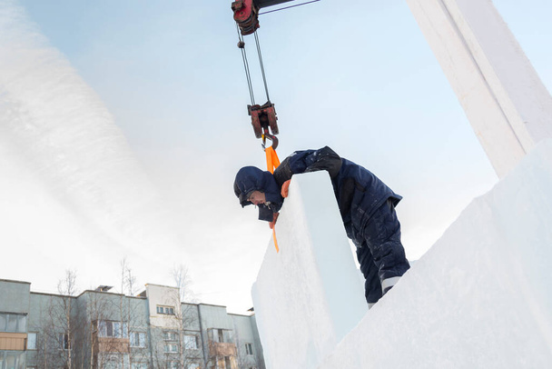 Portret van een arbeider in een blauw jasje met een kap aan de haak van een vrachtwagenkraan bij de bouw van een ijsstad - Foto, afbeelding
