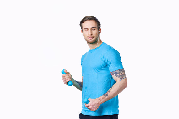 мужчина делает упражнения с гантелями на светлом фоне в голубой футболке и штанах - Фото, изображение