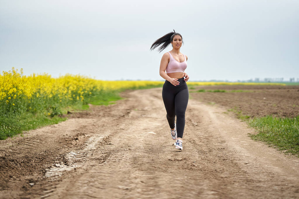 Plus size beautiful latin woman jogging on a dirt road by a canola field - Photo, image