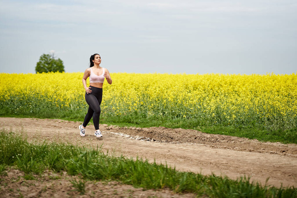 Plus size beautiful latin woman jogging on a dirt road by a canola field - Φωτογραφία, εικόνα