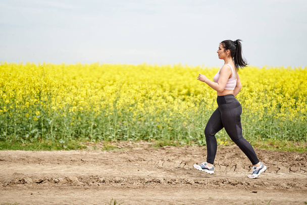 Plus size beautiful latin woman jogging on a dirt road by a canola field - Photo, Image