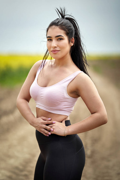 Plus size female runner posing in running outfit on a dirt road by a canola field in the countryside - Foto, afbeelding
