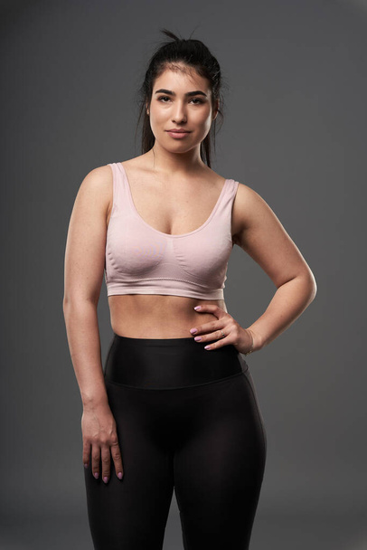 Happy plus size hispanic young woman in fitness clothing posing against gray background - Foto, afbeelding