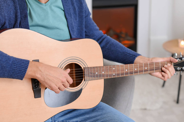 Young man playing guitar near fireplace at home - Foto, Imagen