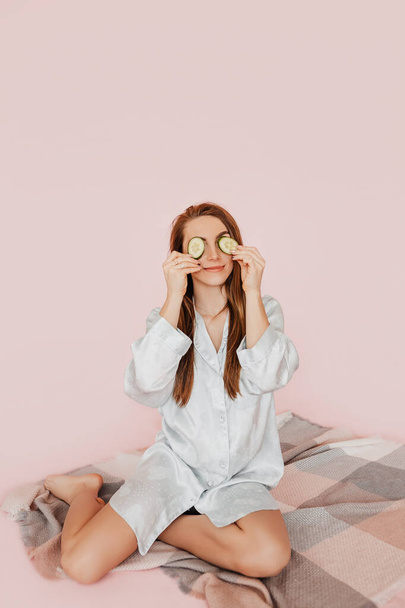Girl make homemade face beauty masks. Cucumbers for the freshness of the skin around the eyes. Woman take care of youthful skin. Model laughing and having fun in spa on pink background - Zdjęcie, obraz