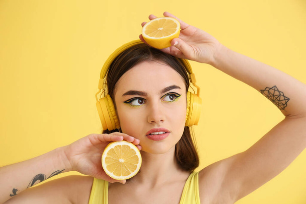 Κομψή νεαρή γυναίκα με ακουστικά και λεμόνι σε κίτρινο φόντο - Φωτογραφία, εικόνα
