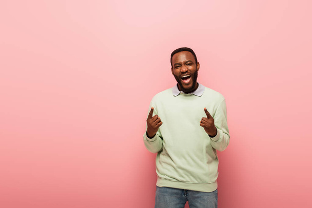 Positive african american man pointing with fingers on pink background  - Фото, зображення