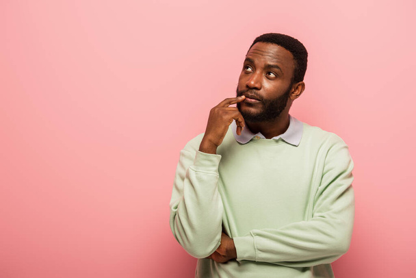 Réfléchi homme afro-américain avec doigt près des lèvres regardant loin isolé sur rose  - Photo, image