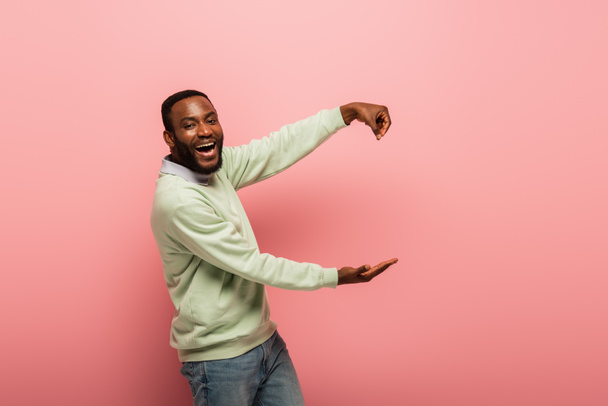 Positive african american man pointing with hand and holding something on pink background  - Фото, изображение