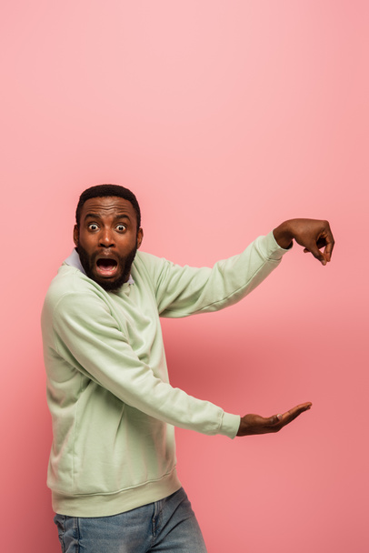 Amazed african american man holding something and pointing isolated on pink  - Fotoğraf, Görsel