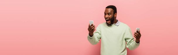 Hombre afroamericano positivo con teléfonos inteligentes sobre fondo rosa, bandera  - Foto, imagen