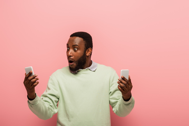 Amazed african american man holding cellphones on pink background  - Foto, afbeelding