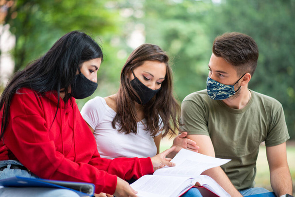 Three students studying together sitting on a bench outdoor and wearing masks during coronavirus times - Foto, imagen